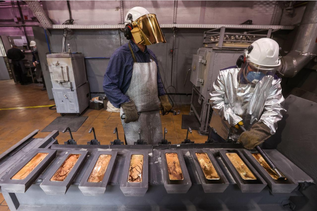 Workers wearing protective equipment handling metal bars inside a precious-metals processing facility.