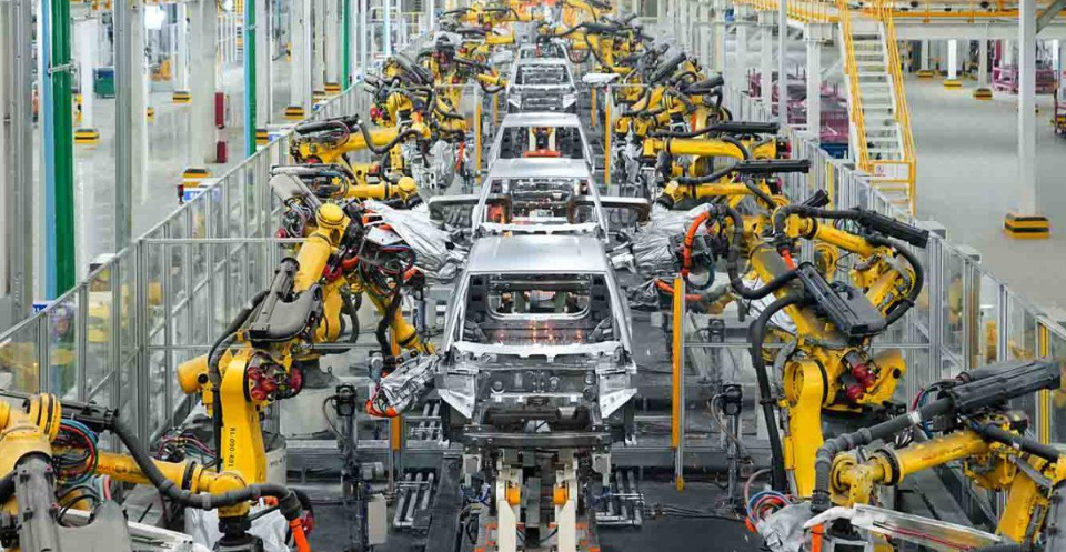 Cars moving along an automotive assembly line surrounded by industrial robotic arms.