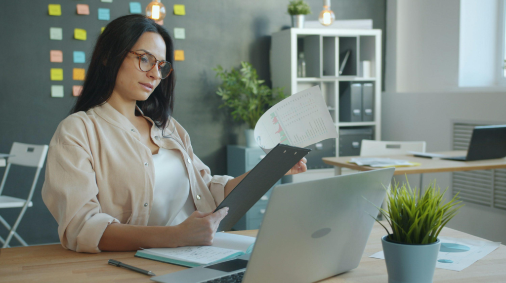 Business professional reviewing documents beside a laptop at an office desk.