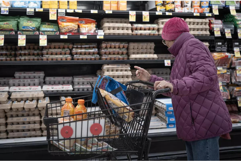 A shopper wearing a purple jacket stands beside a grocery cart in front of refrigerated egg shelves at a supermarket.